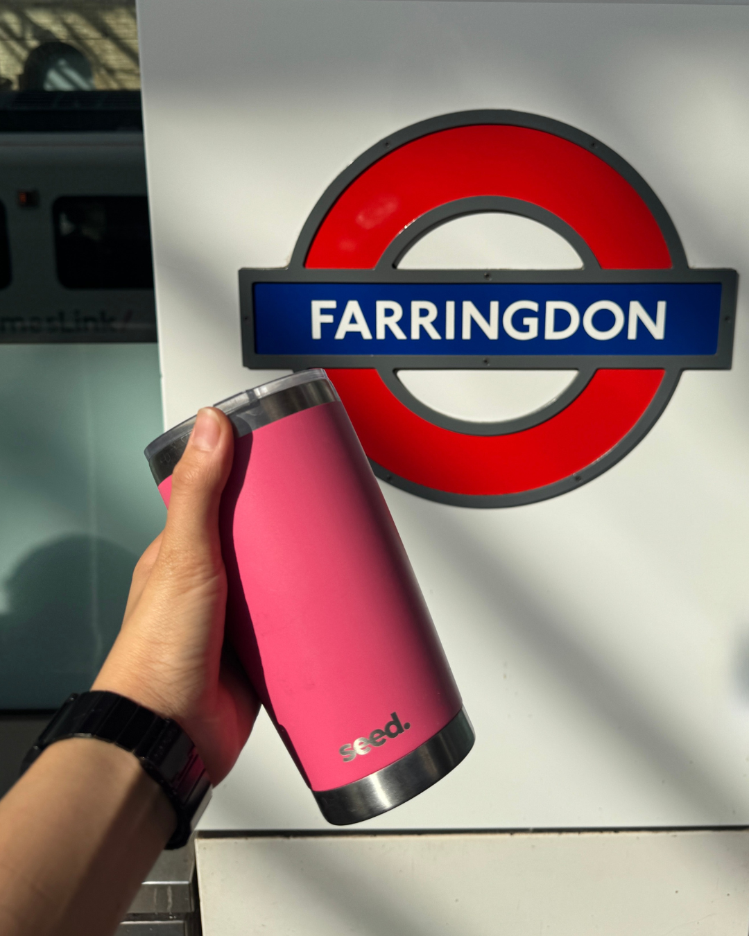 Pink insulated bottle held by a hand in front of a underground railway in London, Farringdon sign.  insulated stainless steel travel coffee mug 600mL candy pink