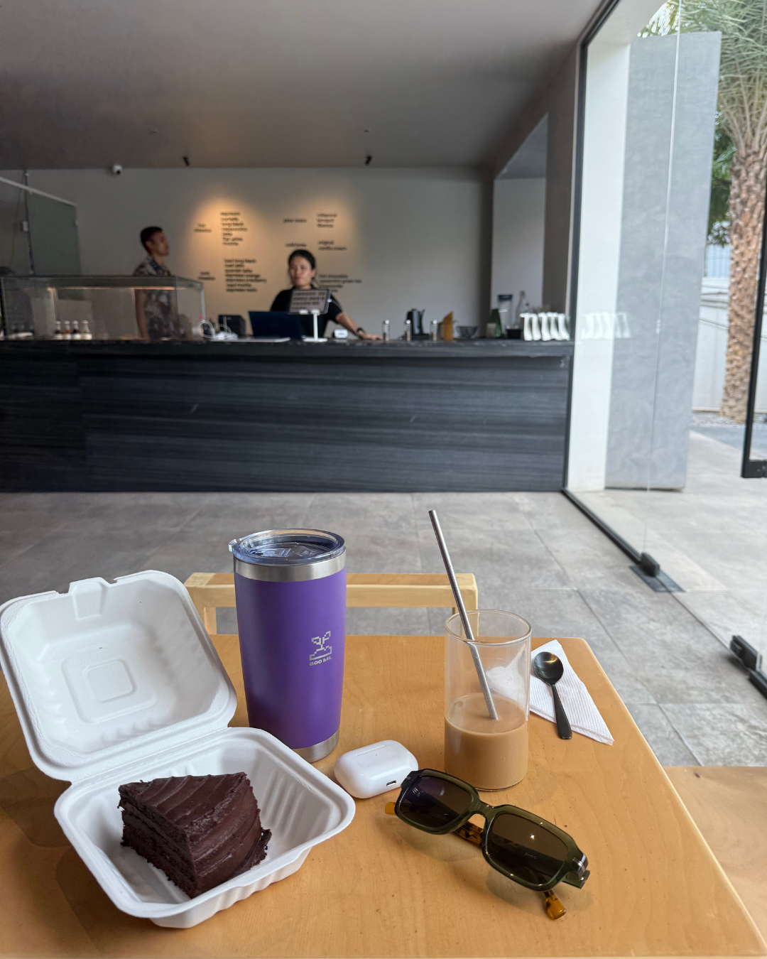 Table with a purple travel coffee mug with coffee in it, chocolate cake, and sunglasses, airpods in front of a cafe counter. insulated stainless steel travel coffee mug 600mL lavender