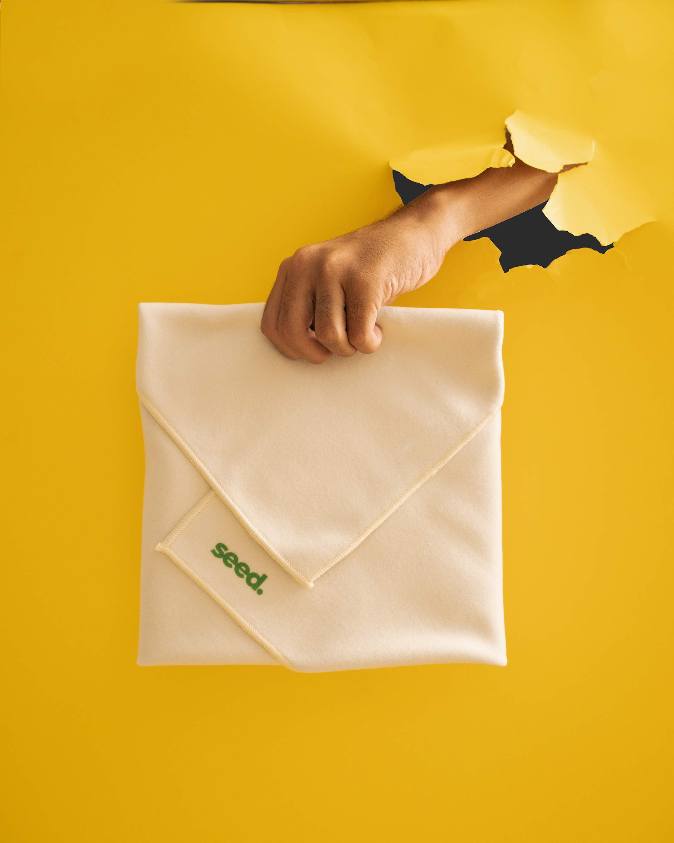 Hand holding a folded beige cloth with 'seed' logo on a yellow background Hold Up Magic Self Adhesive Wrap  Organiser Cat In The Bag