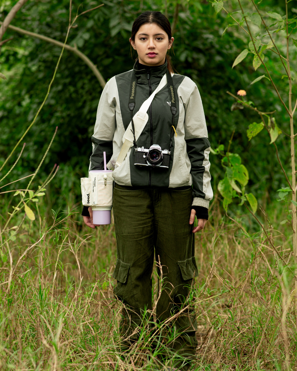 A person standing in a natural outdoor setting with tall grasses and trees in the background, wearing a gray and black utility sling bag  sleeve with an adjustable strap, a lilac stainless steel tumbler, and carrying a camera. milkyway & lilac
