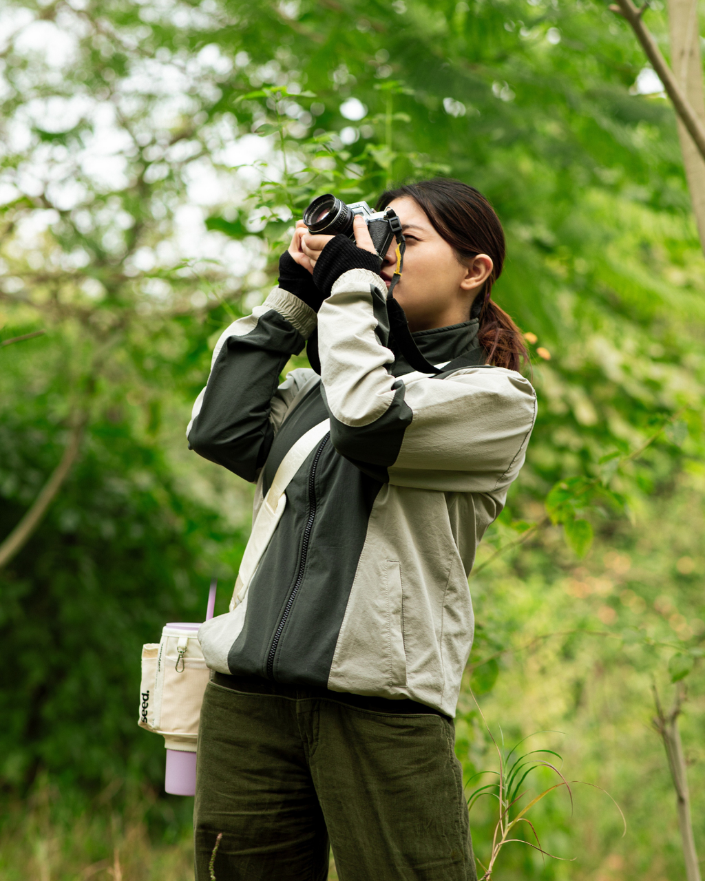 A person is taking pictures on a film camera a lilac stainless steel tumbler with a white sleeve slung across her shoulder. The setting appears to be outdoors with greenery in the background. seed move carry all sling multi-purpose utility sleeve bag for stainless steel tumblers milkyway & lilac