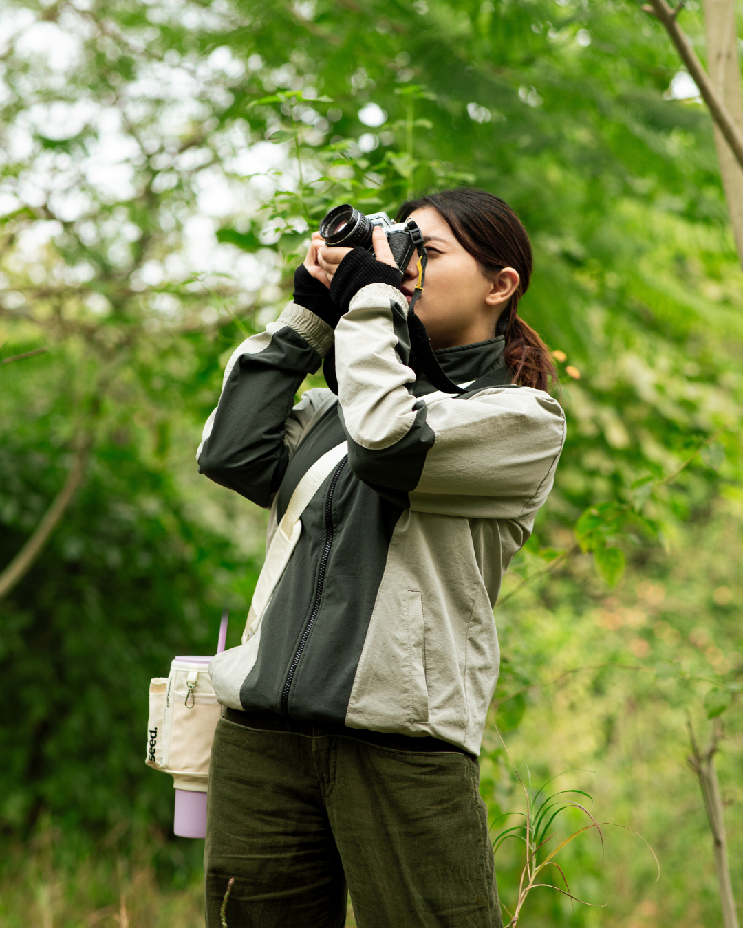 A person is taking pictures on a film camera a lilac stainless steel tumbler with a white sleeve slung across her shoulder. The setting appears to be outdoors with greenery in the background. seed move carry all sling multi-purpose utility sleeve bag for stainless steel tumblers milkyway