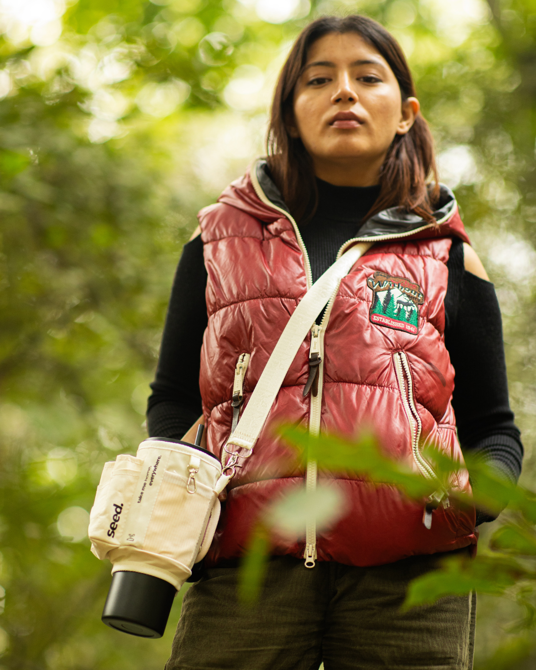 A person wearing a red puffer vest and black top, holding a beige and black tumbler in a outdoor setting with green foliage in the background. milkyway dark knight
