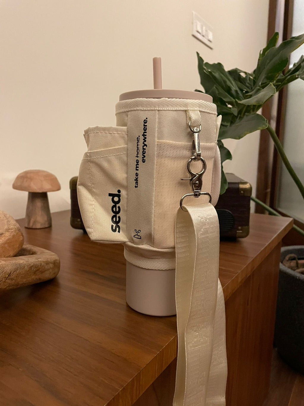 Beige insulated stainless steel tumbler water bottle with matching utility sleeve sling bag  on a wooden surface with plants in the background milkyway & milk tea