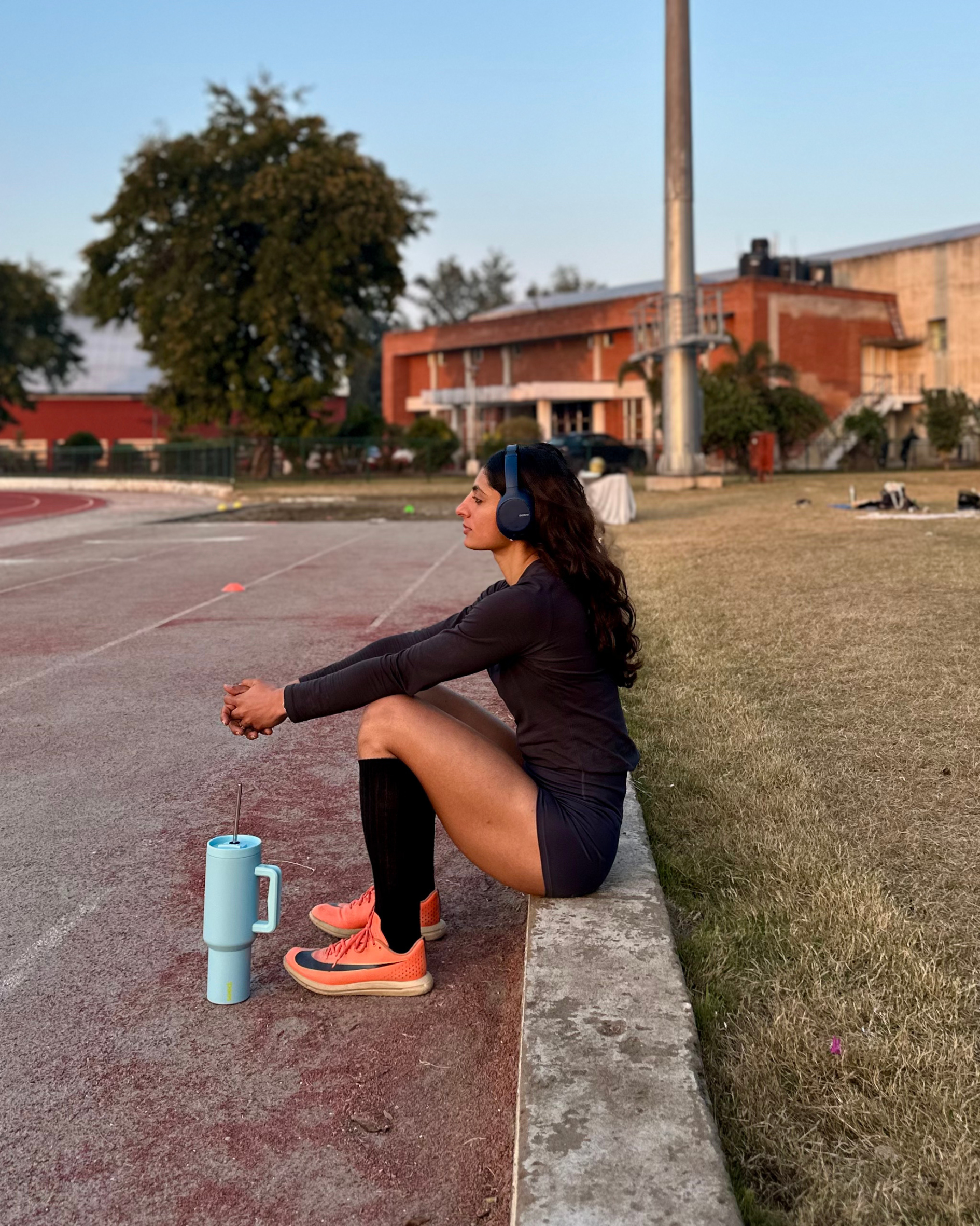 Person sitting on a track with a water bottle and headphones, wearing athletic gear. stainless steel tumbler 1.2L baby blue 